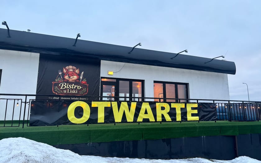 Budynek bistro od strony drogi: biała elewacja, czarny baner z logo lisa i żółty napis OTWARTE